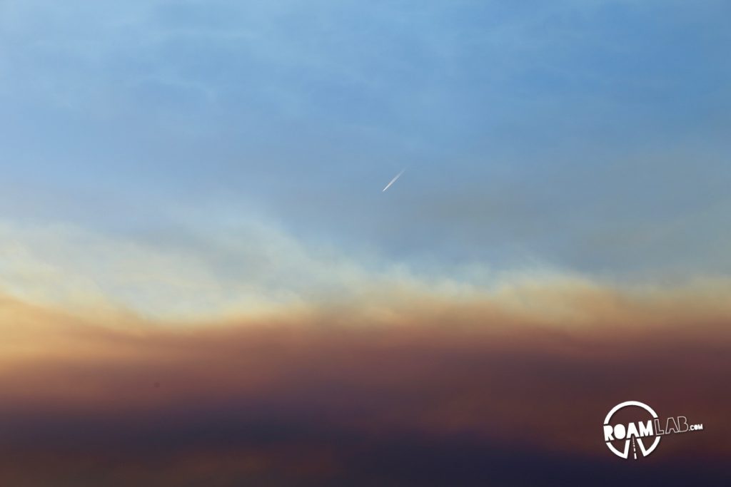 A jet trail descending behind an ephemeral plane of smoke from a nearby fire.