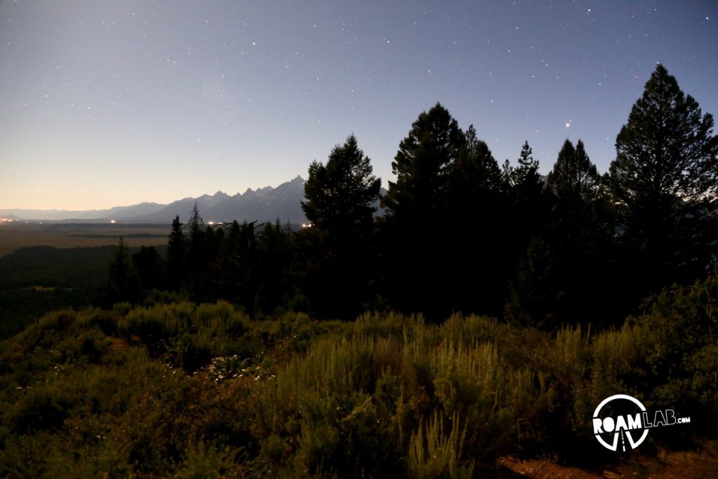 The Grand Tetons peak out behind the trees and brush of Signal Mountain. A nearly full moon casts a gradient across the sky washing out the stars from the left.