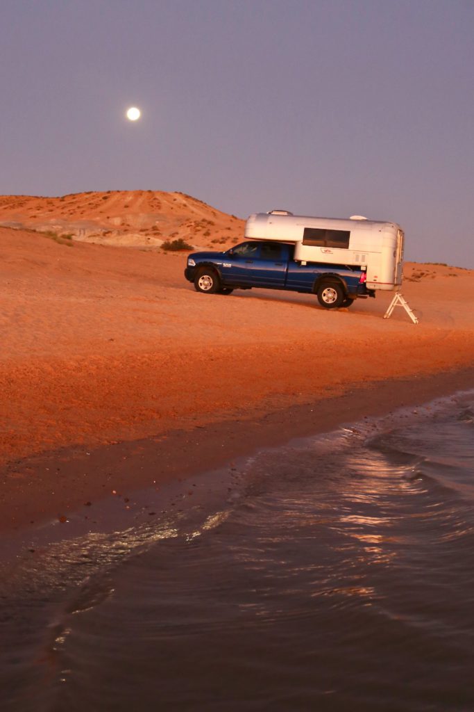 Vintage truck camper parked on a beach with the moon setting behind a sand dune.