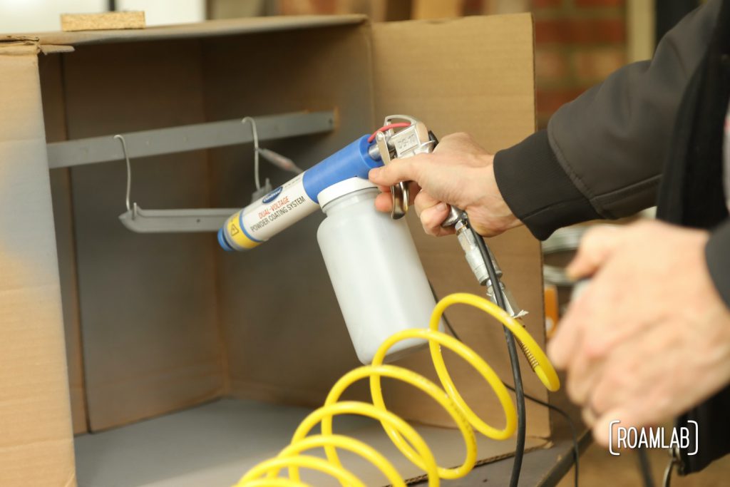 Closeup of hands holding a powder coating gun to apply on an emblem in the backgroun