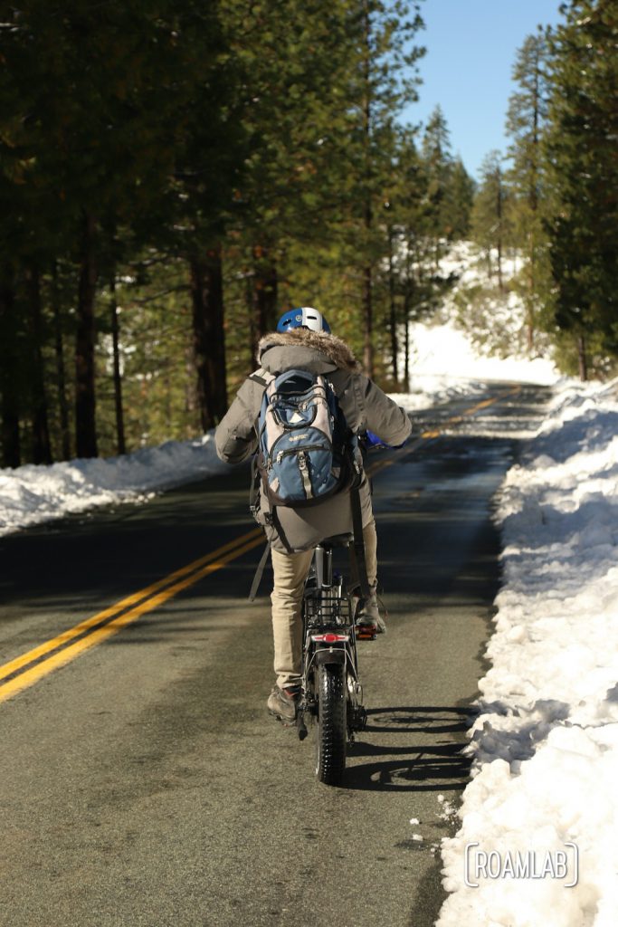 Man with a blue backpack riding a bike down a paved road with snow on each side in Foresthill OHV Trail System.