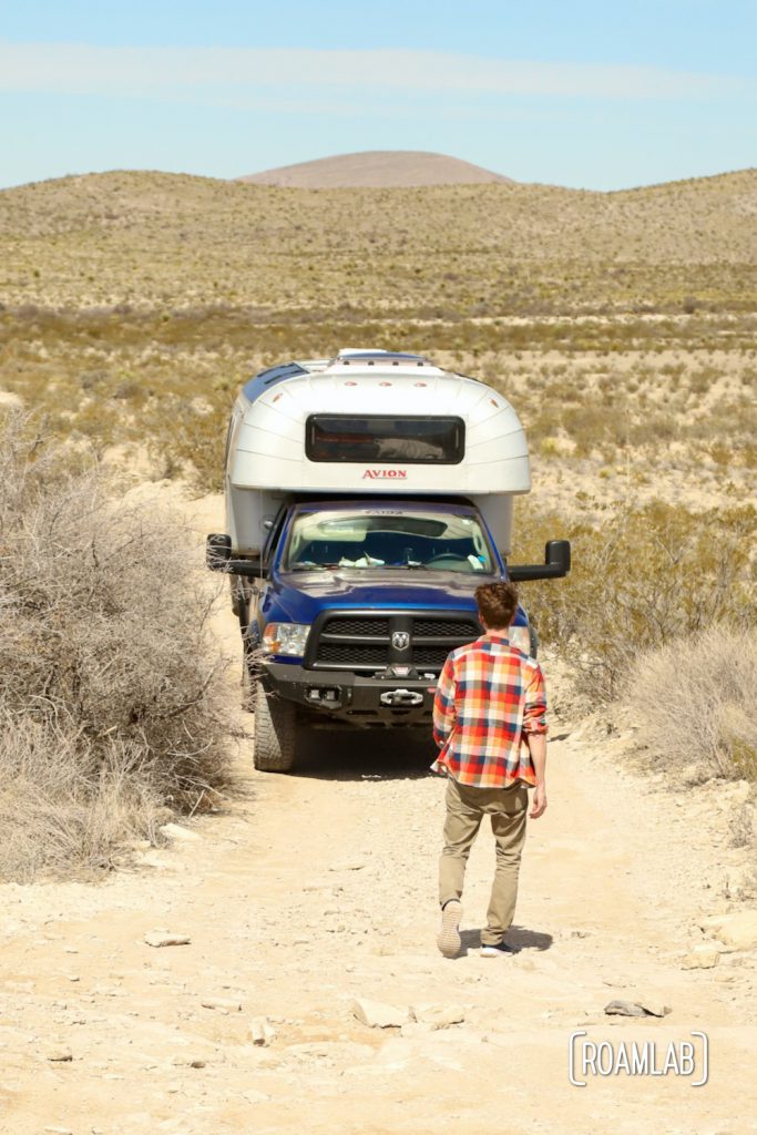 Man in a plaid shirt inspecting a rocky dirt road in front of a dusty 1970 Avion C11 truck camper along Old Ore Road in Big Bend National Park, Texas.