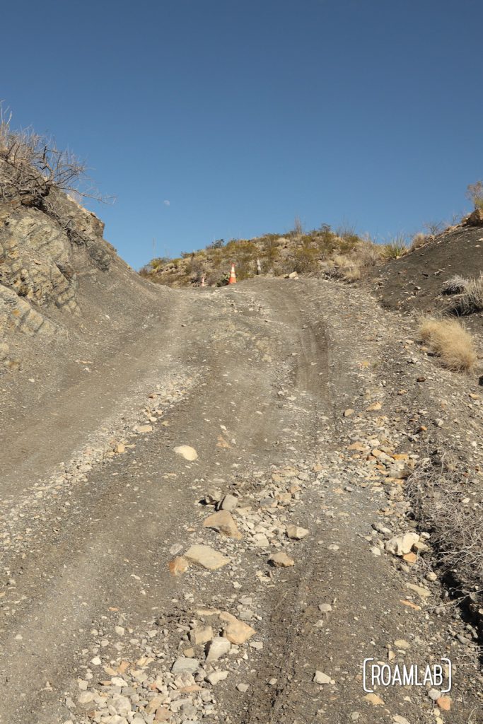 A steep rocky rutted road with a safety cone at the top and blue sky above along Old Ore Road in Big Bend National Park, Texas.