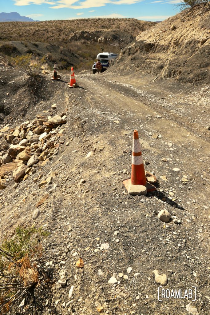 Orange safety cones marking a crumbling edge of dirt desert road with a 1970 Avion C11 truck camper approaching along Old Ore Road in Big Bend National Park, Texas.