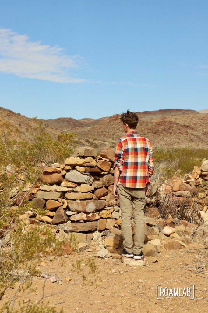 Man in a colorful plaid shirt standing next to stacked stone walls of a crumbling structure in the desert along Old Ore Road in Big Bend National Park, Texas.