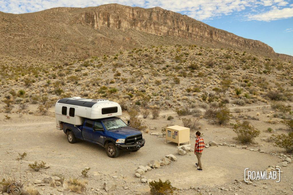 Man walking towards a 1970 Avion C11 truck camper with cliffs in the background parked at the Telephone Canyon 1 Campsite off Old Ore Road in Big Bend National Park.