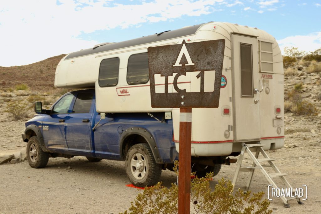 "TC 1" sign with a tent icon cut into rusting sheet metal on a pole in front of a silver 1970 Avion C11 truck camper on a blue truck with desert hills and blue sky in the background off Old Ore Road in Big Bend National Park, Texas.