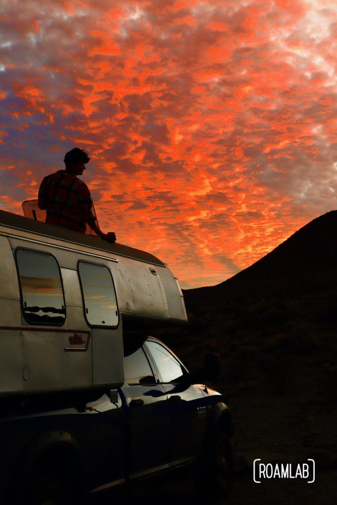 Brilliant red sunset lighting up the clouds silhouetting a man peaking out of a roof hatch of a 1970 Avion C11 truck camper parked at Telephone Canyon 1 campsite off of Old Ore Road in Big Bend National Park, Texas.