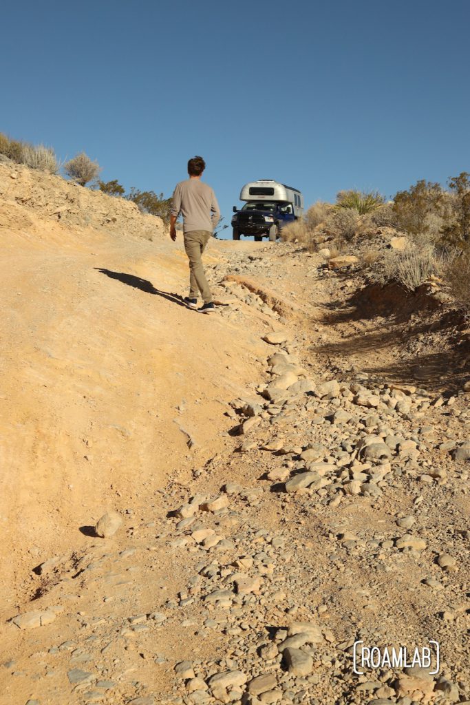 Man scouting the decent along a steep and rocky dirt road with a 1970 Avion C11 truck camper waiting along the Old Ore Road in Big Bend National Park, Texas