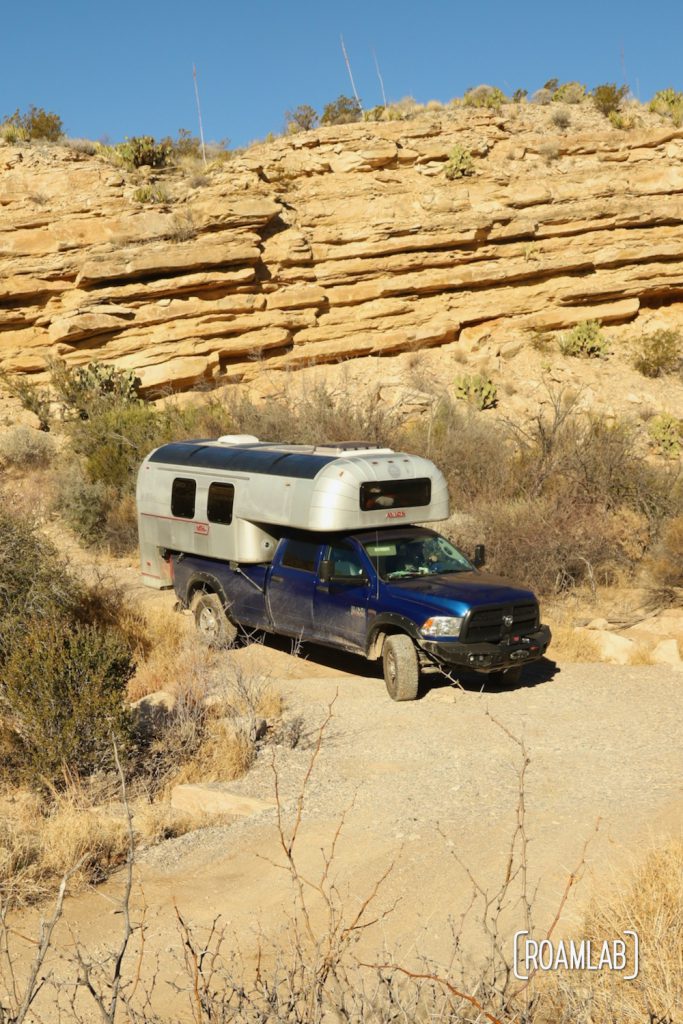 Dusty 1970 Avion C11 truck camper rounding a corner on a dusty road through a wash with yellow ridged cliffs.