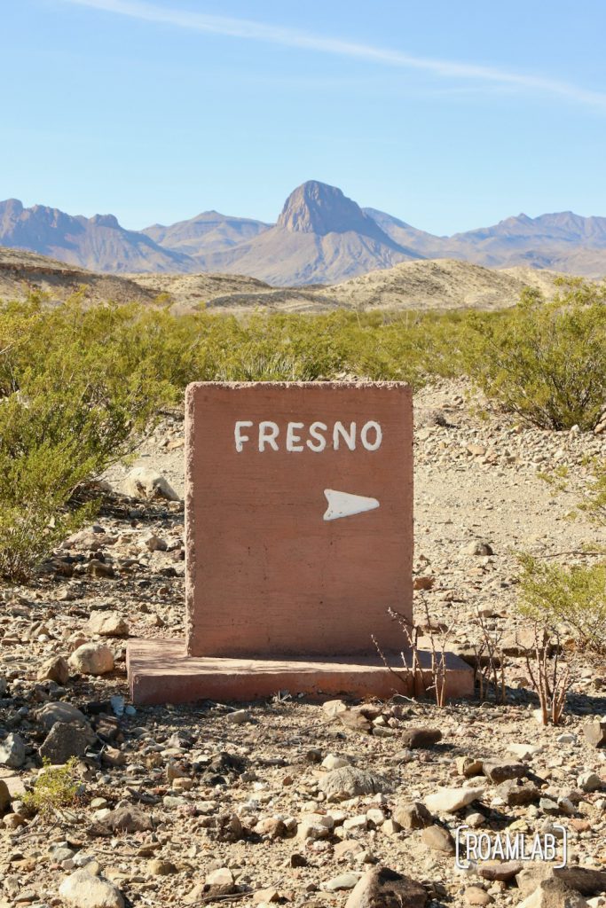 Pinkish stone marker pointing right to Fresno Campground, among rocky desert with shrub brush and mountains in the distance off River Road in Big Bend National Park, Texas.