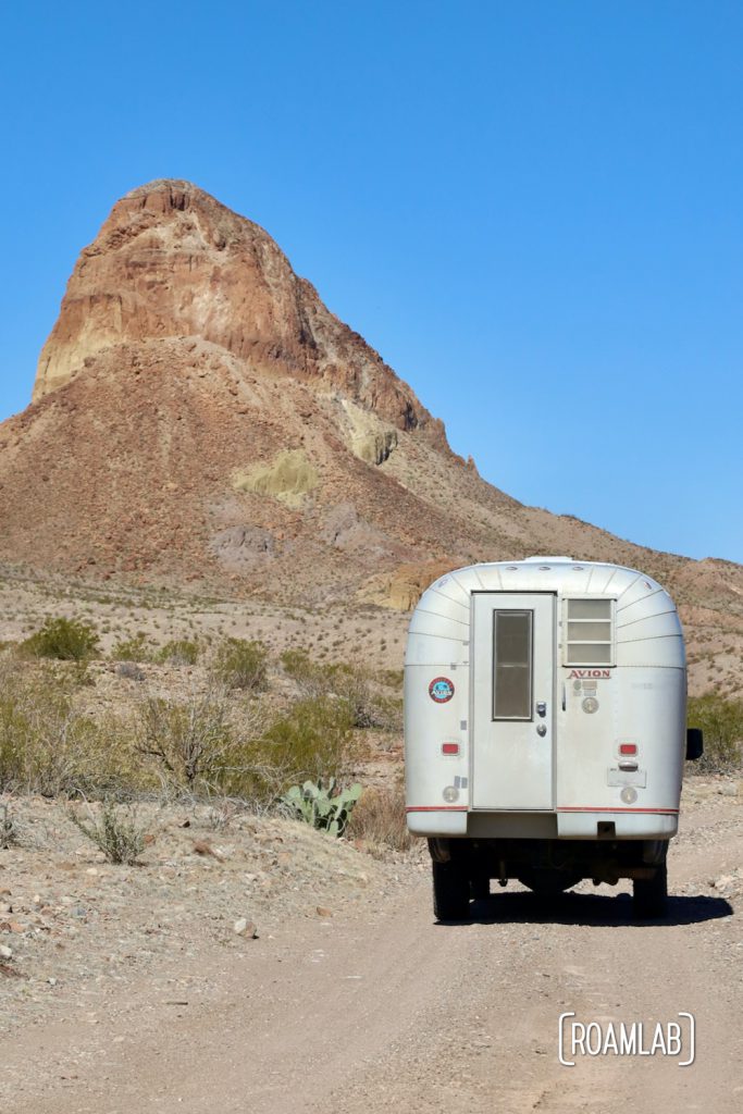 Silver 1970 Avion C11 truck camper on a dirt road with a large colorful bluff to the left at the western terminus of River Road in Big Bend National Park, Texas.