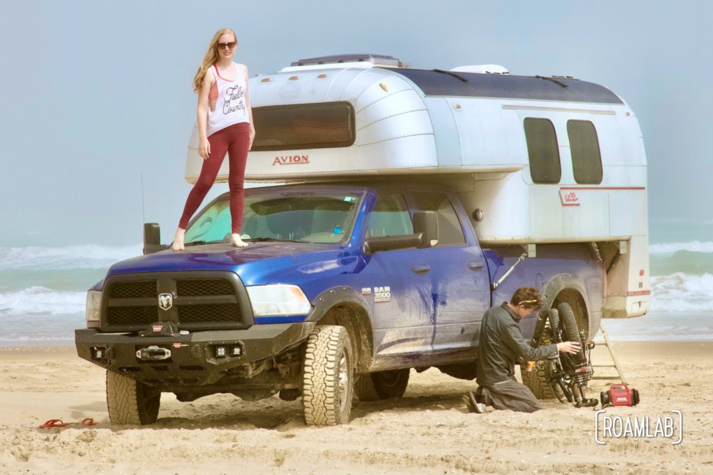 Woman standing on the hood of a blue truck with a 1970 Avion C11 truck camper and a man kneeling on the beach while tuning up a bicycle on South Padre Island, Texas.