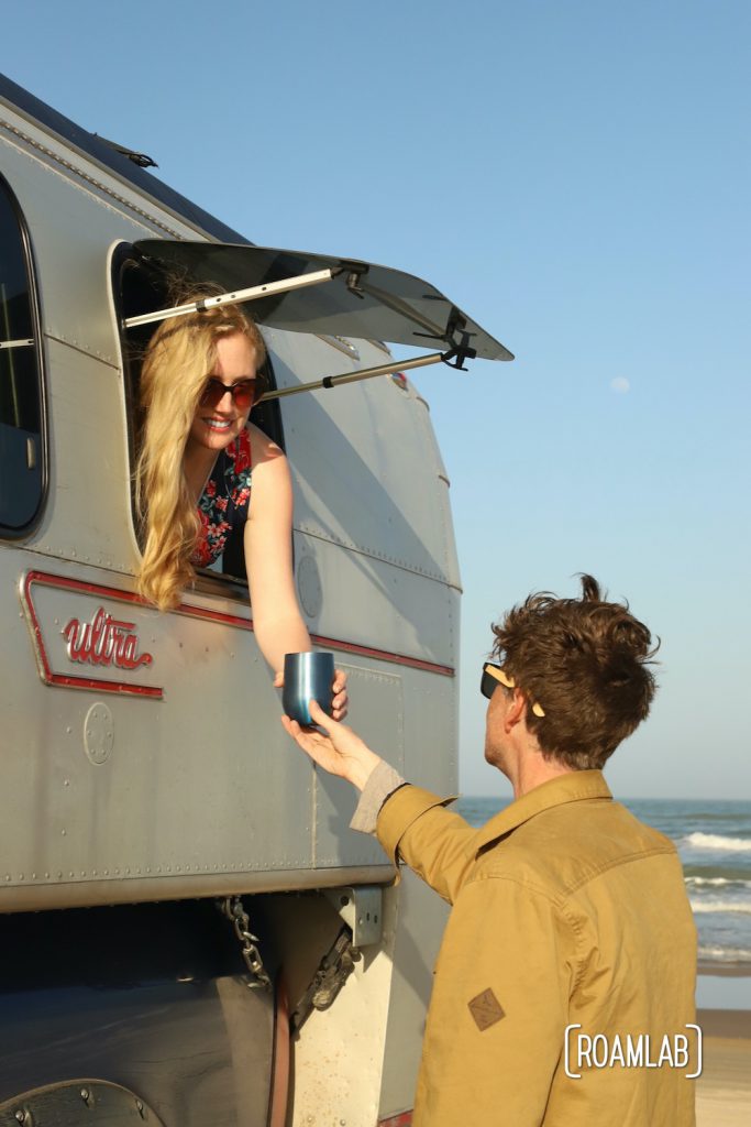 Woman in a 1970 Avion C11 truck camper passing a wine glass to a man standing on the beach with waves in the background in South Padre Island, Texas.