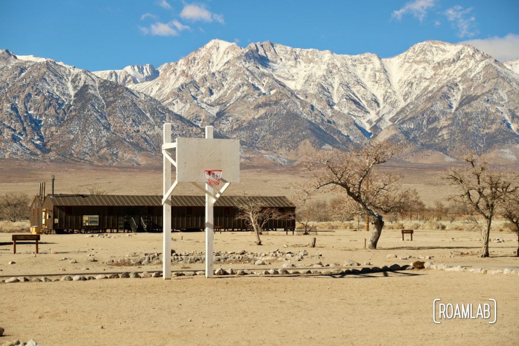 White plywood basket ball hoop over a dirt court with brown barracks in the background and snow capped mountains in the background of Manzanar National Historic Site.