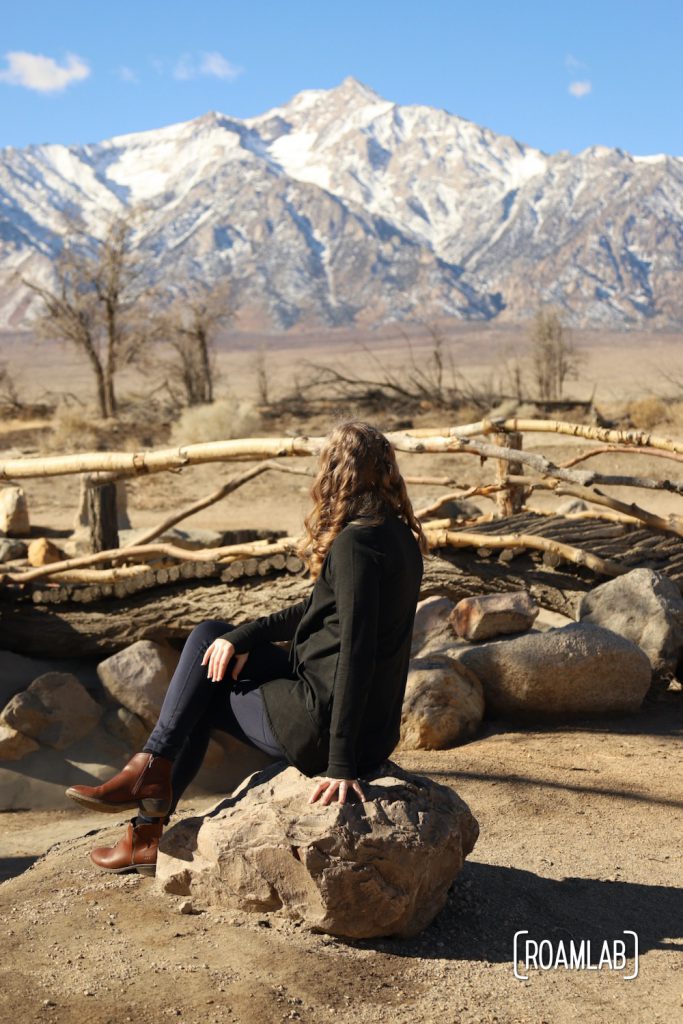 Woman sitting on a stone looking at a rough wooden bridge in Manzanar's Pleasure Park with the Sierra Nevada in the background.