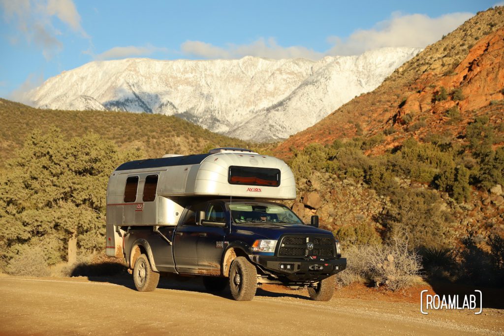 1970 Avion C11 truck camper driving down the red dirt Forest Road 032 in Dixie National Forest with snow capped mountains in the background.