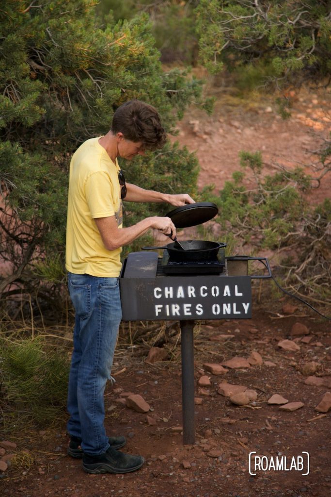 Man looking into a cast iron skillet on top of a grill with "Charcoal fires only" spray painted on the side.