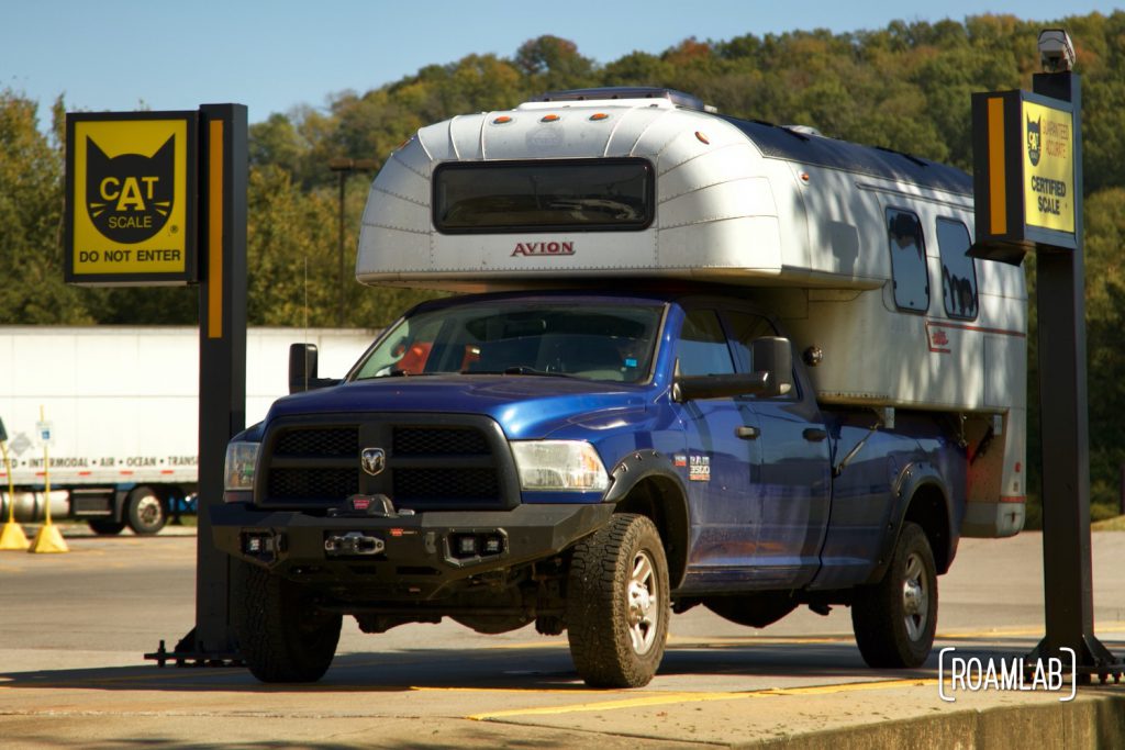Vintage silver aluminum 1970 Avion C11 truck camper on a blue 2015 Ram 3500 truck parked on CAT scale pad with fall color forest hill in the background.