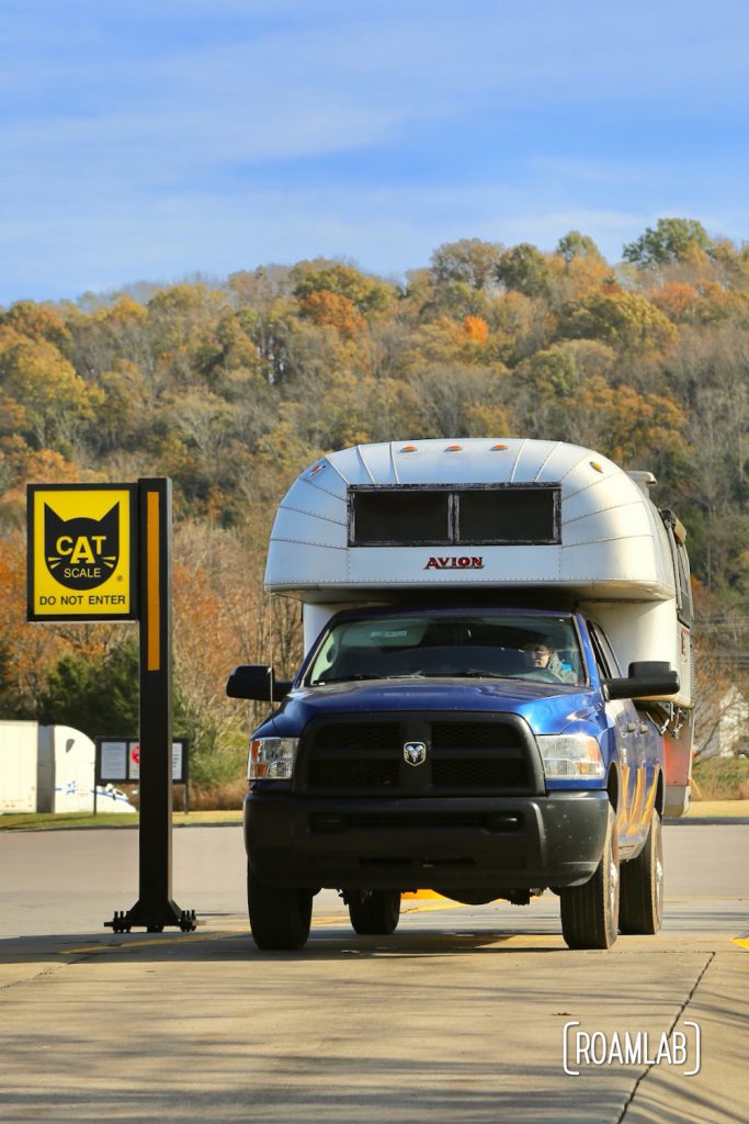 Silver 1970 Avion C11 truck camper sitting on a blue 2015 Ram 3500 with yellow CAT Scales sign to the left and colorful fall hillside in background.