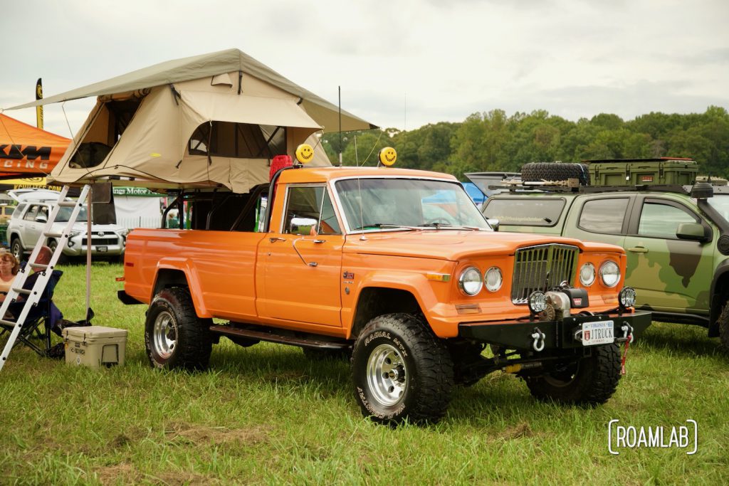 Bright orange Jeep J20 truck with roof top tent camped in a grassy field at Overland Expo East 2021 in Arrington, Virginia.