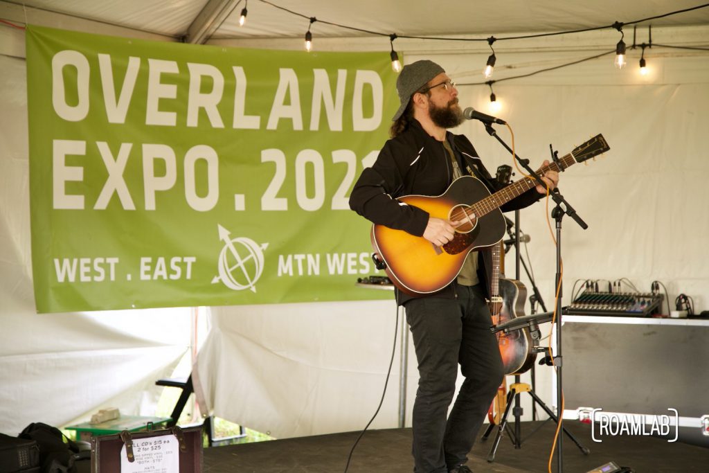 Man strumming guitar and singing into microphone in front of poster for Overland Expo, 2021.