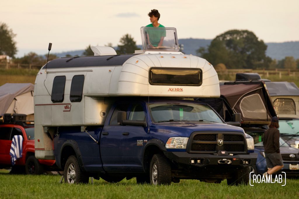 Man looking out of a roof hatch of a 1970 Avion C11 truck camper parked among other rigs at Overland Expo East 2021 in Arrington, Virginia.