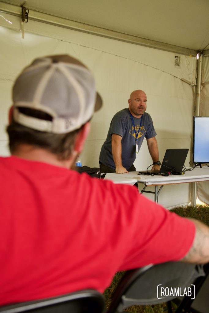 Looking over the shoulder of a man in a red shirt watching a man lecturing at Overland Expo East 2021 in Arrington, Virginia.