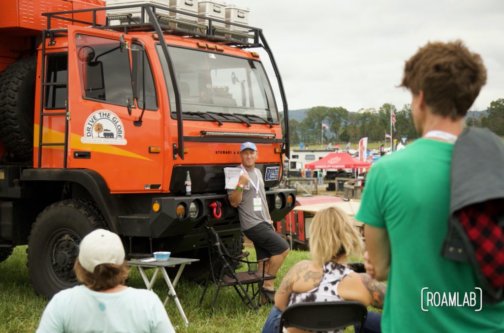 Man presenting massive orange rig to a group of seated onlookers at Overland Expo East 2021 in Arrington, Virginia.