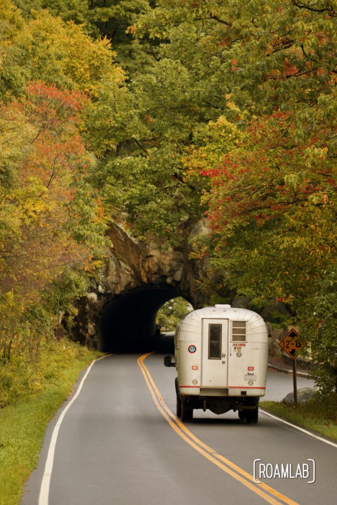 Silver 1970 Avion C11 truck camper approaching a tunnel with colorful foliage.