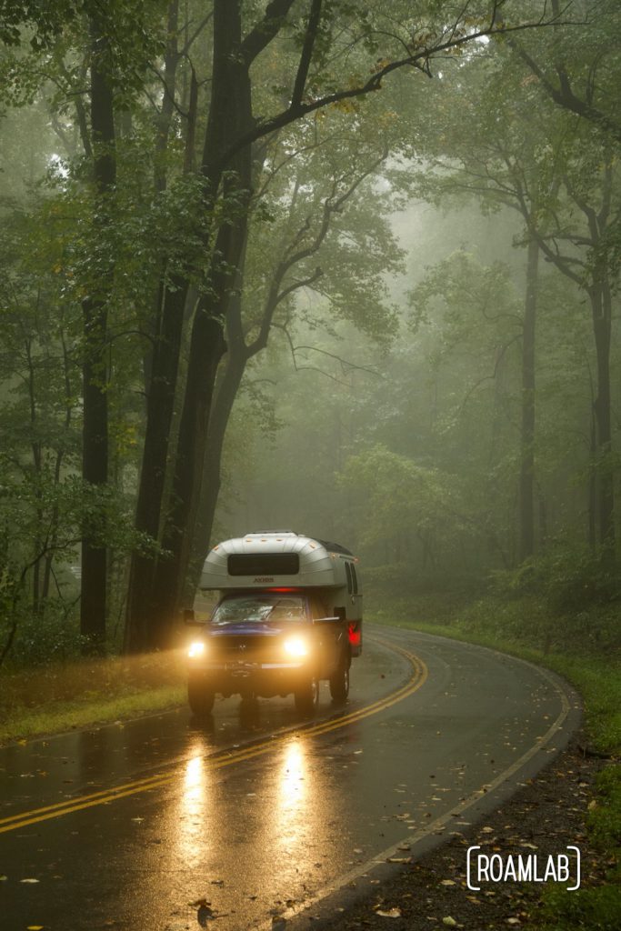 1970 Avion C11 truck camper with headlights on driving through a misty forest on a wet road in Shenandoah National Park.