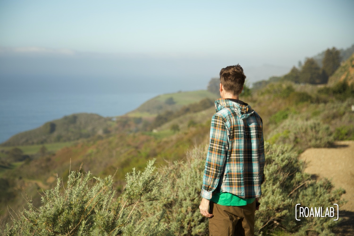Man in plaid jacket standing on a green hillside looking away from the camera, towards the ocean on the horizon in Big Sur, California.