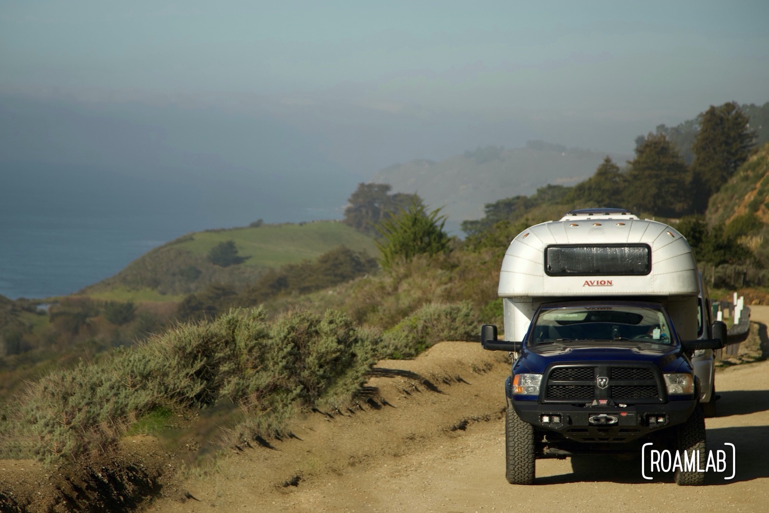 1970 Avion C11 truck camper on a blue Ram truck parked on a dirt pullout on a green hillside with the Pacific ocean in the background in Big Sur, California along Highway 1.