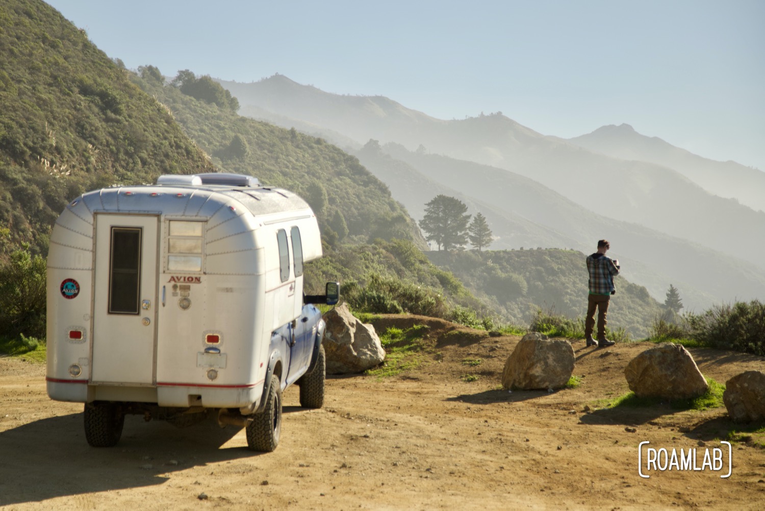 Man standing on a cliff side, taking a picture next to a 1970 Avion C11 truck camper along Highway 1 in Big Sur, California.