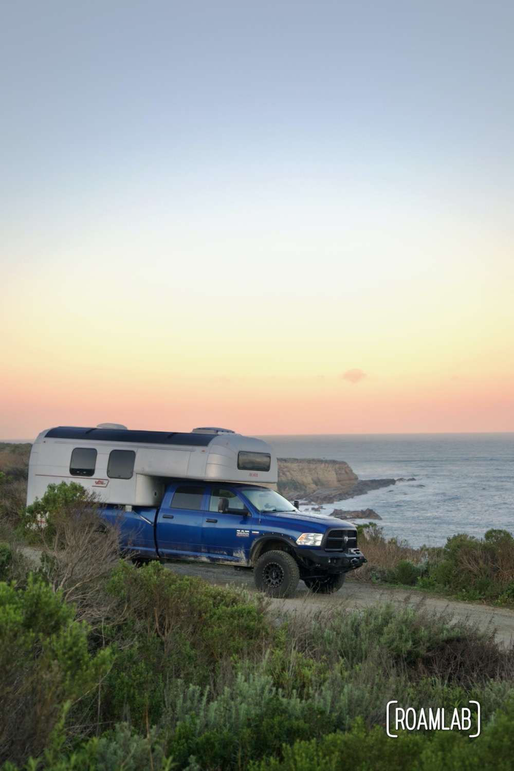 1970 Avion C11 truck camper on a paved road with the Montaña de Oro State Park coastline at sunrise in the background.