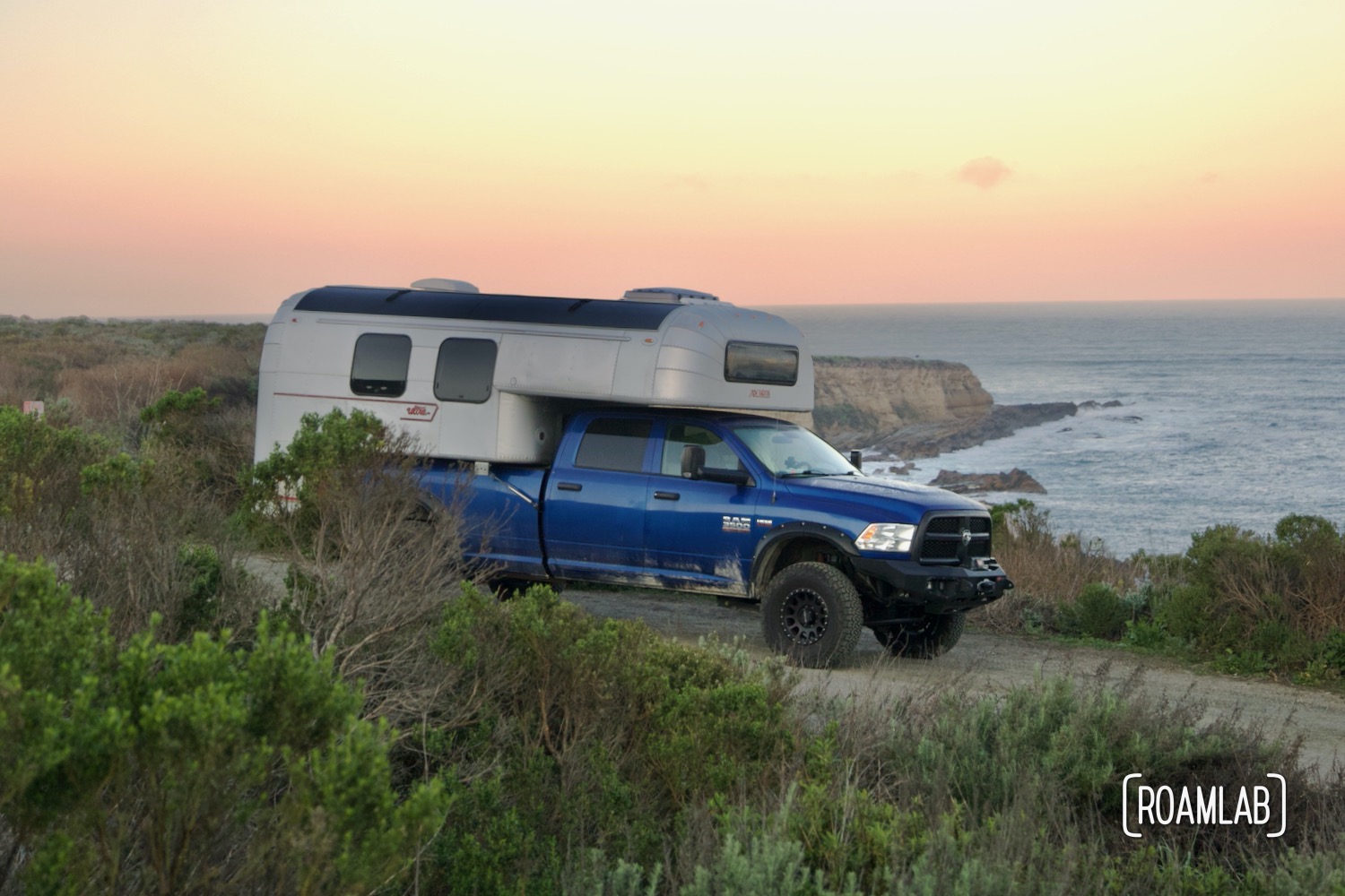1970 Avion C11 truck camper on a paved road with the Montaña de Oro State Park coastline at sunrise in the background.