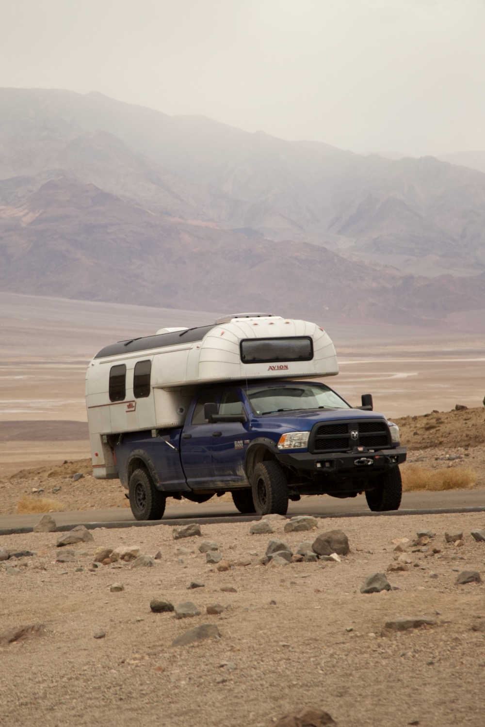 1970 Avion C11 truck camper driving up a road in Death Valley National Park with partially obscured mountains in the background.