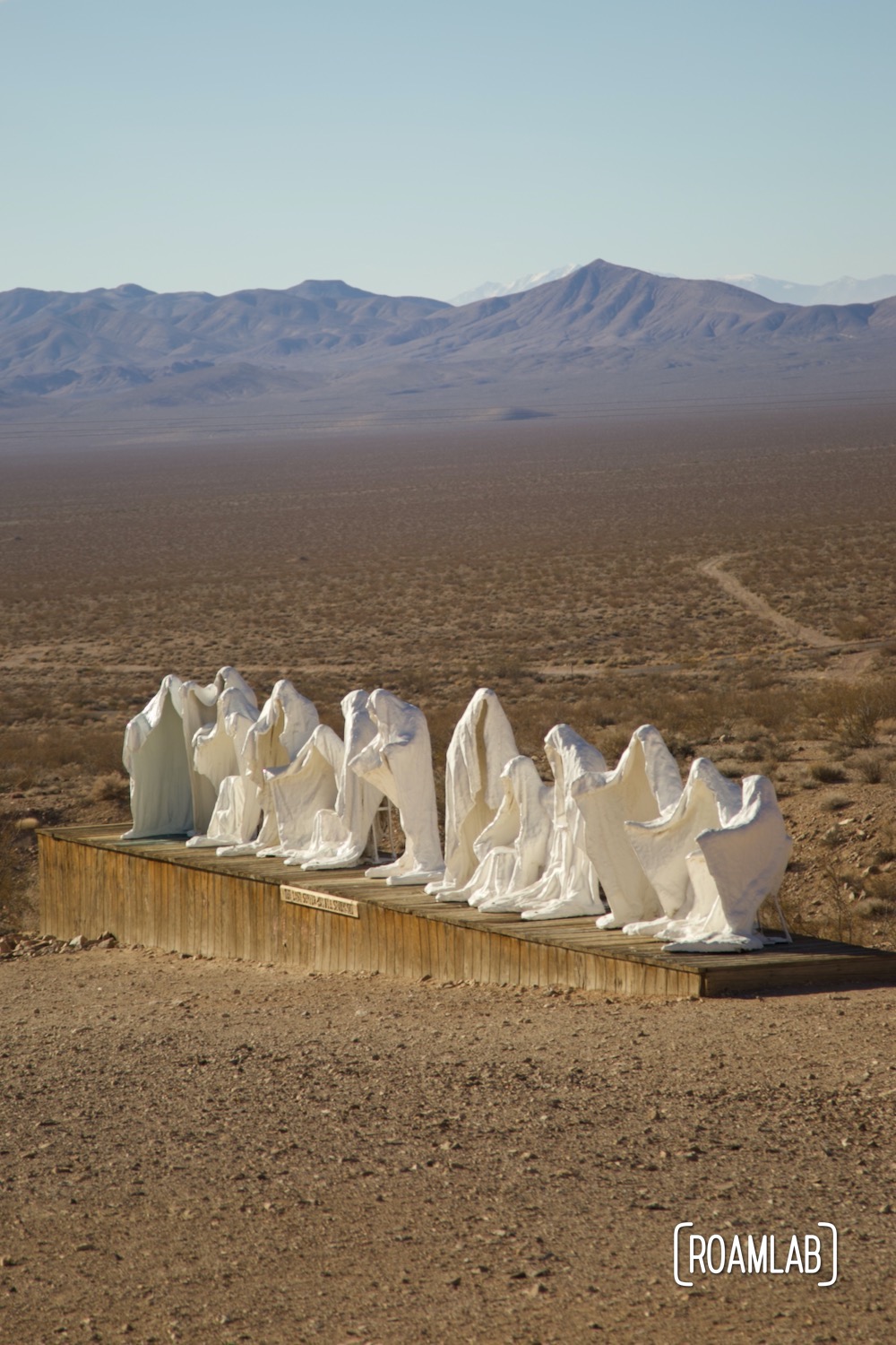 Plaster ghost sculptures in the form of the last supper on a wood platform with the Nevada desert and mountains in the background.