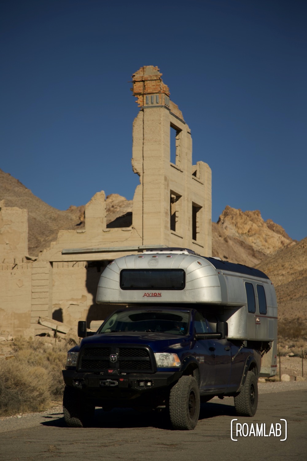 1970 Avion C11 truck camper driving by a collection of ruined buildings with desert mountains rising in the background.