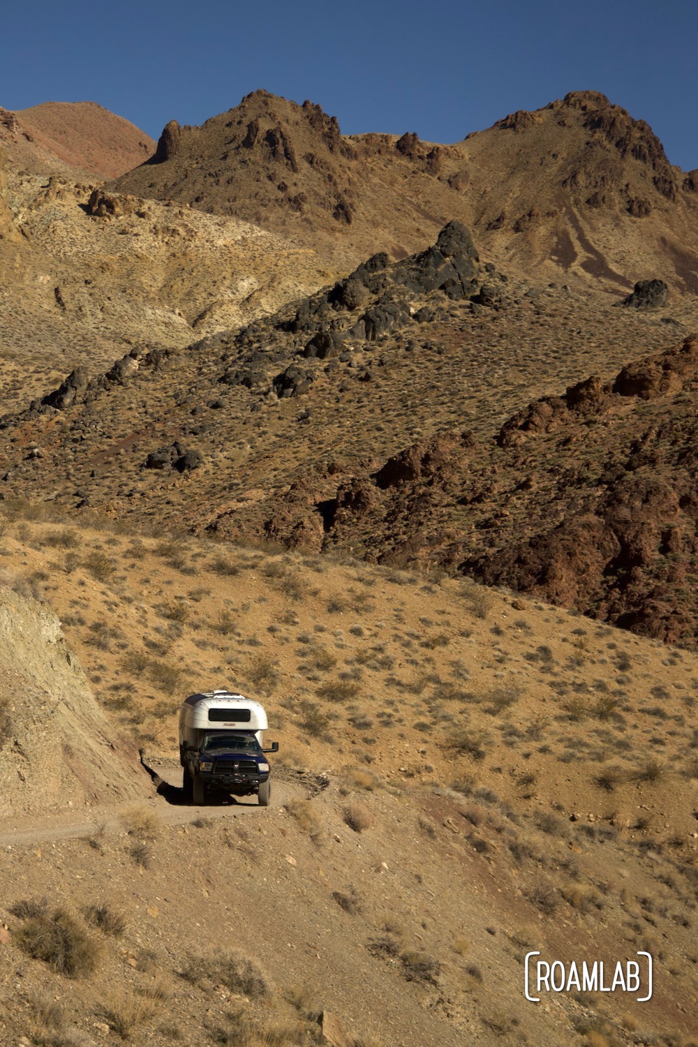 Road dropping away with mountains in the background of a 1970 Avion C11 truck camper following the dirt Titus Canyon Road in Death Valley National Park, California.