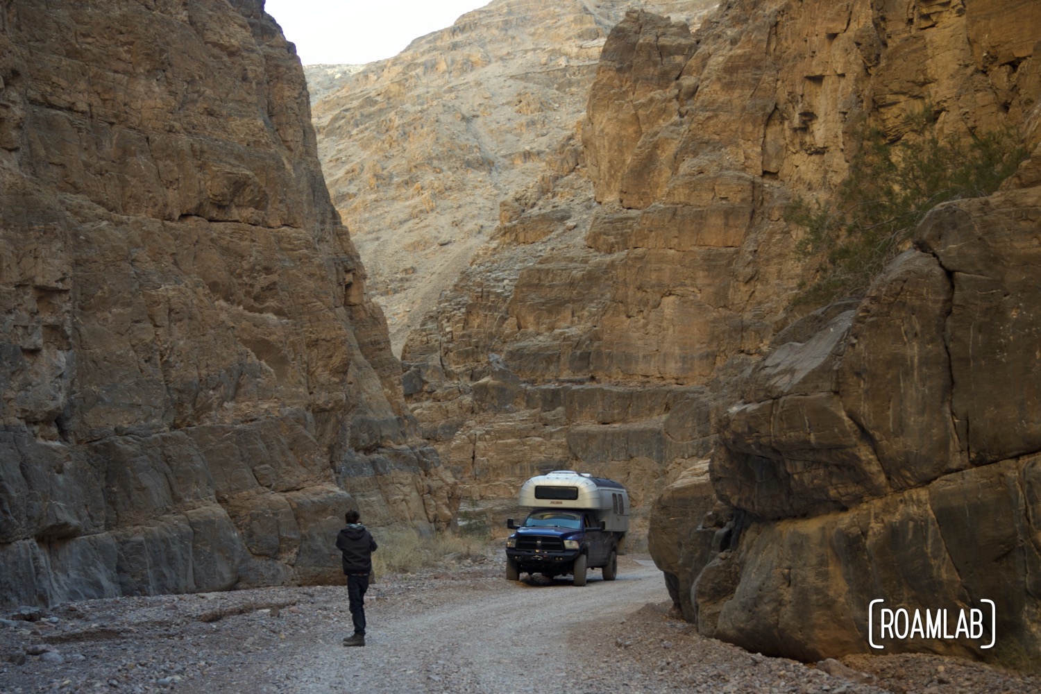 Man Pausing on a gravel wall to photograph a 1970 Avion C11 truck camper following the dirt Titus Canyon Road in Death Valley National Park, California.