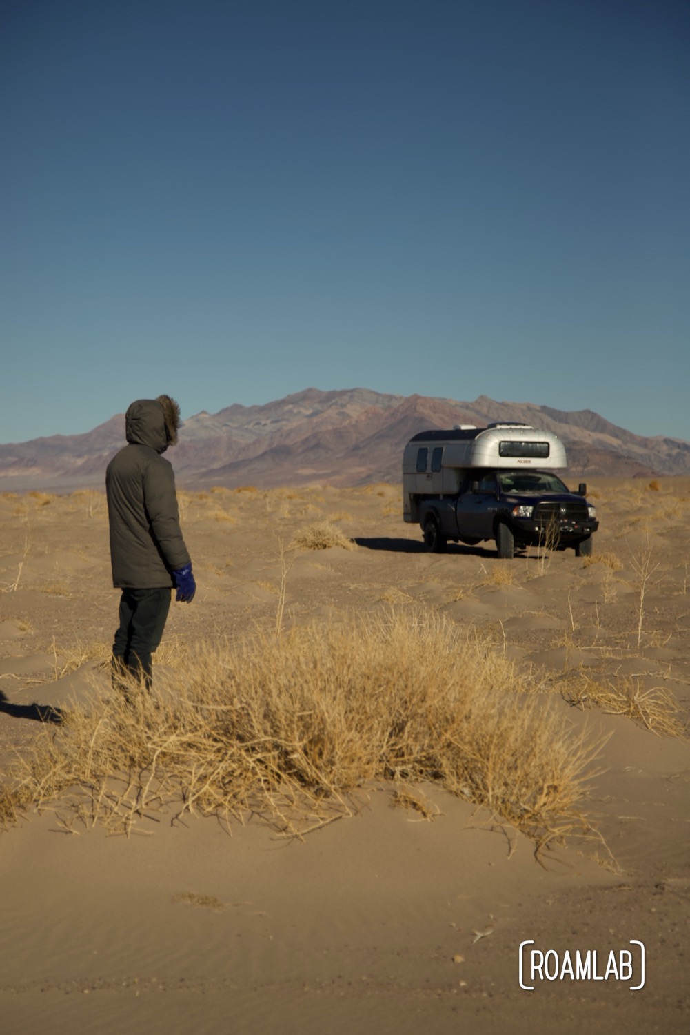 Man walking around a 1970 Avion C11 truck camper parked in the sand in front of distant mountains in Big Dune Recreational Area.