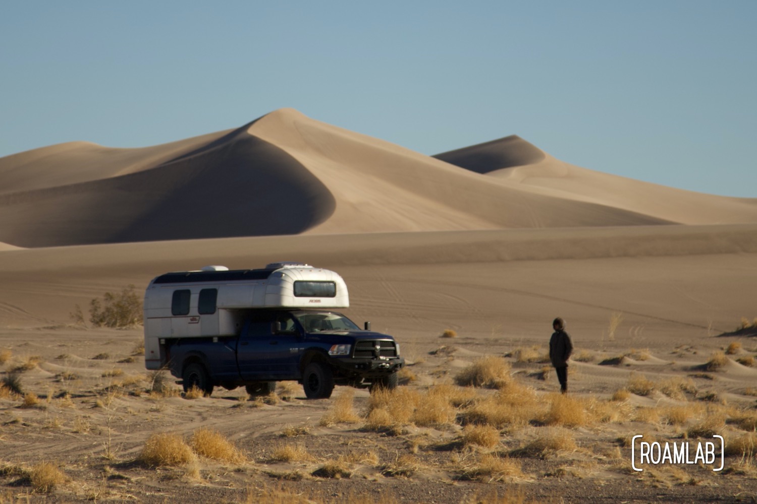 Man walking around a 1970 Avion C11 truck camper parked in the sand in front of the dunes of Big Dune Recreational Area.