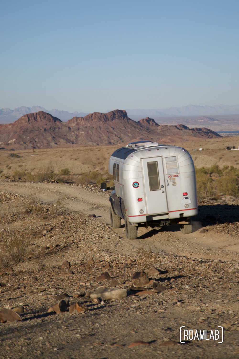 1970 Avion C11 truck camper driving along a dirt road with mountains and Lake Havasu in the background.