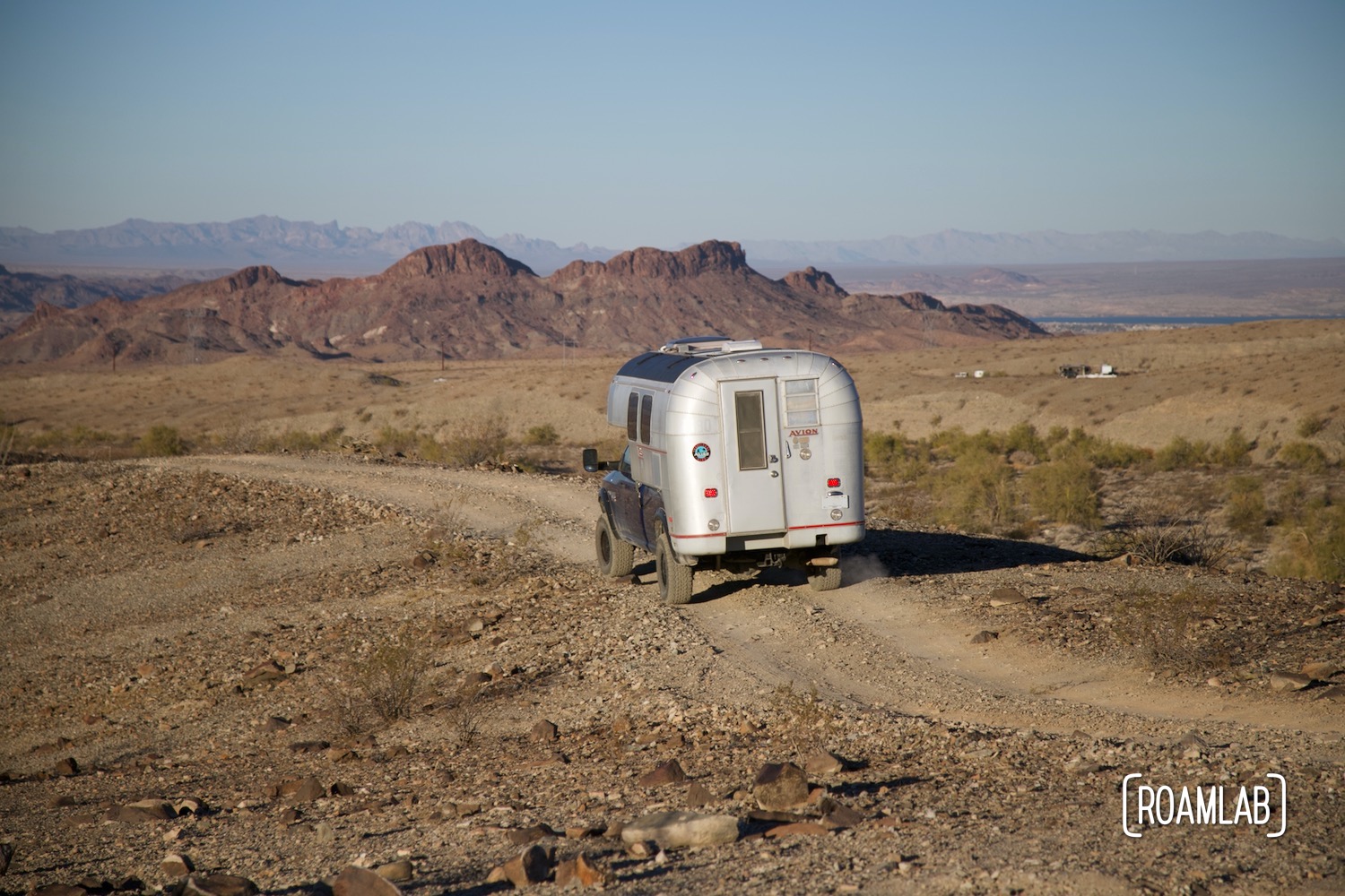 1970 Avion C11 truck camper driving along a dirt road with mountains and Lake Havasu in the background.
