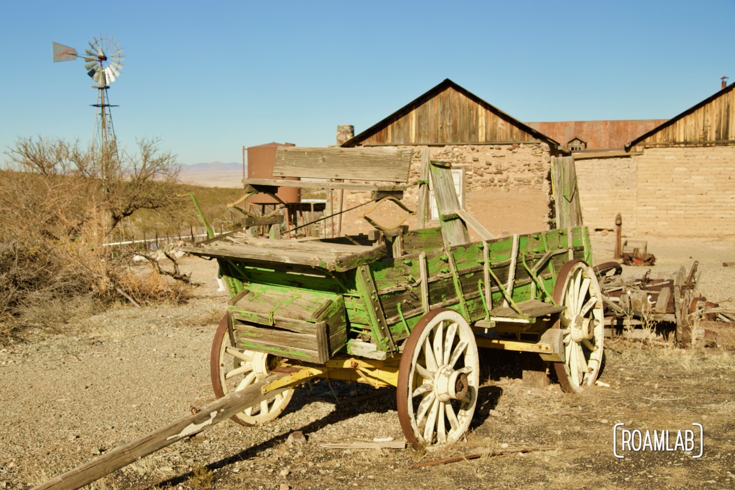 Green wagon in front of an aging adobe and wood structure with a windmill in the background in Shakespeare Ghost Town, New Mexico.