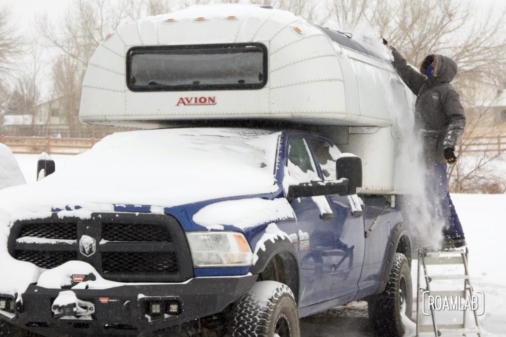 Man on a step ladder brushing snow off solar panels mounted on a 1970 Avion C11 truck camper.