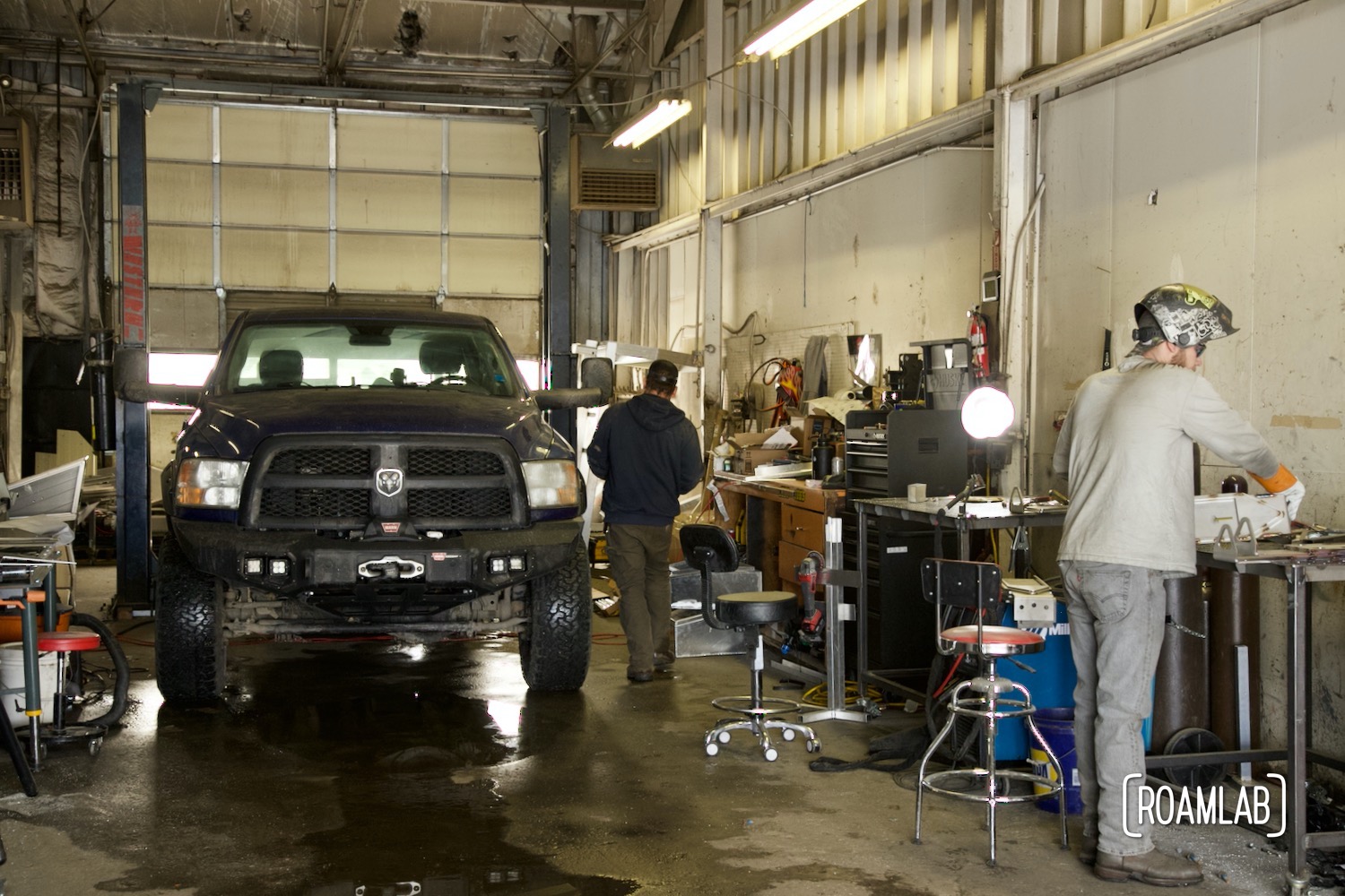 Man walking around truck parked in an industrial garage while other man standing at desk with welding helmet.