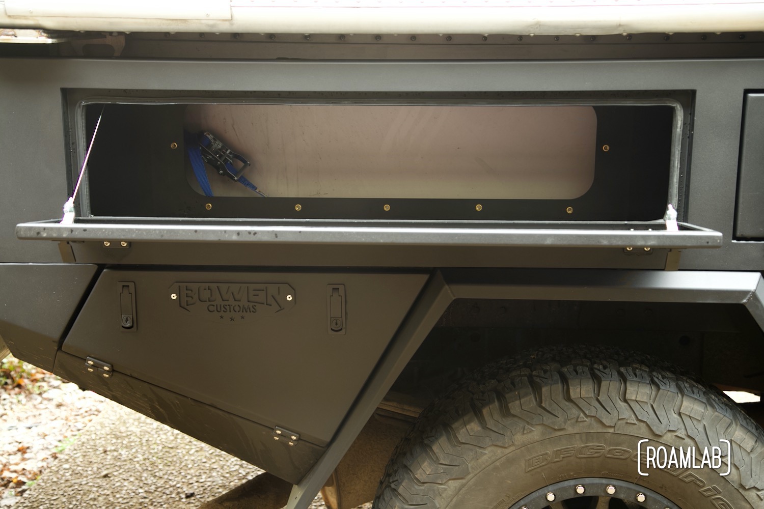 Open storage box with inner wall removed, exposing a ratchet strap securing the camper to the Bowen Customs aluminum truck bed.