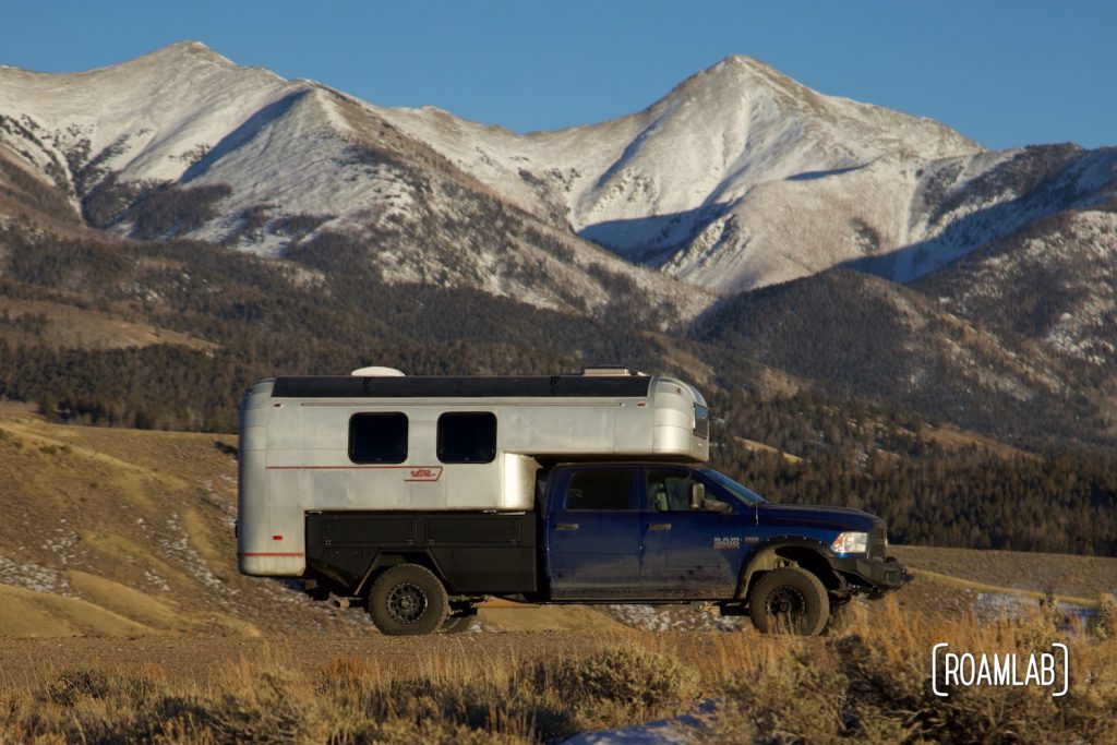 Silver aluminum 1970 Avion C11 truck camper on a blue Ram truck and black Bowen Customs truck camper in front of the Rocky Mountains.